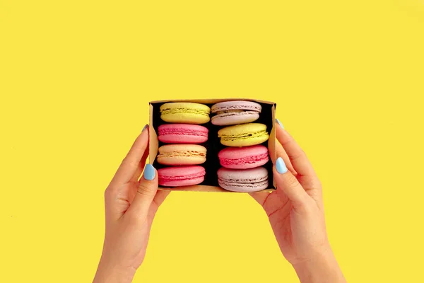 Kadınların elleri, sarı arka planda renkli badem kurabiyeleriyle dolu bir kutunun içinde. — Stok fotoğraf