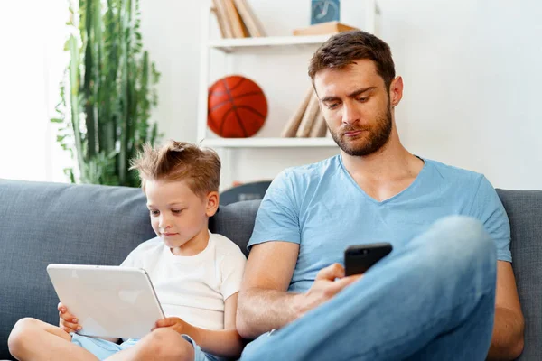 Pequeño niño usando tableta digital y su padre usando teléfono inteligente sentarse juntos en el sofá — Foto de Stock