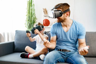 Baba oğul kanepede oturup VR gözlükleriyle oyun oynuyorlar.