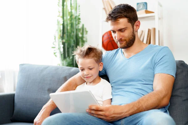 Pequeño niño y su padre viendo vídeos en la tableta digital juntos sentados en el sofá — Foto de Stock
