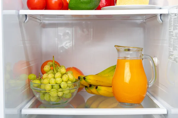 Glass pitcher of orange juice and fruits on fridge shelf — Stock Photo, Image