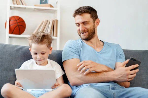 Pequeño niño y su padre viendo vídeos en la tableta digital juntos sentados en el sofá — Foto de Stock