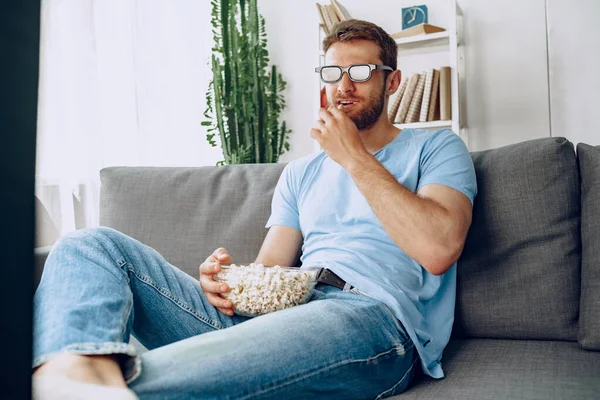 Bearded guy in 3d glasses sitting on sofa and watching movie with popcorn
