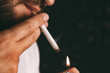 Sakallı adam çakmakla sigara yakıyor.
