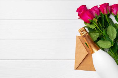 Pembe güller, şampanya şişesi ve beyaz ahşap arka planda bir zarf.