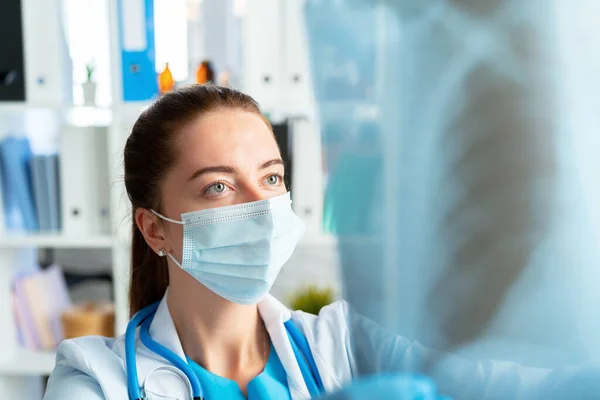 Tıbbi maskeli kadın doktor akciğerlerin röntgen taramasını inceliyor. — Stok fotoğraf