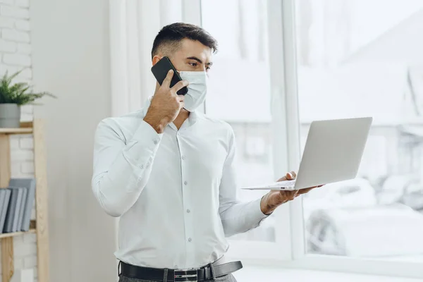 Jóképű férfi üzletember orvosi maszkban használja a laptopját, miközben a mobilján beszél. — Stock Fotó