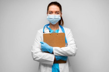 Tıbbi maskeli kadın doktor gri arka planda pano ile ayakta duruyor.