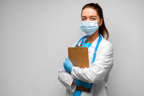 Kobieta lekarz w masce medycznej stojącej ze schowkiem na szarym tle — Zdjęcie stockowe