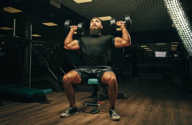 Kaslı genç adam karanlık bir spor salonunda ağırlık kaldırıyor.