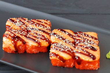 Ahşap masada, siyah tabakta servis edilen fırında suşi rulosu.