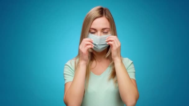 Mujer joven se quita la máscara médica y sonríe contra el fondo azul — Vídeos de Stock