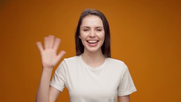 Молода жінка посміхається і трясе рукою, вітаючи когось, на жовтому тлі — стокове відео
