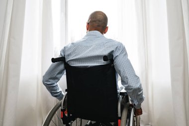 Tekerlekli sandalyedeki Afrikalı Amerikalı adam evde tek başına pencerenin yanında oturuyor.