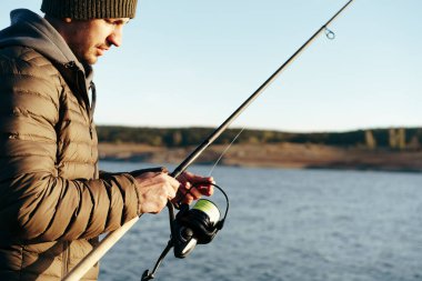 Genç balıkçı elinde oltayla gölün kıyısında duruyor.