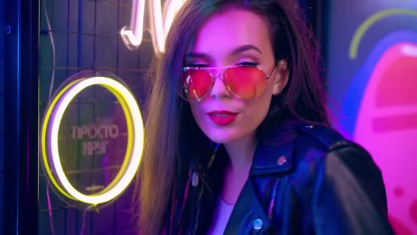 Mixed raced woman with dreadlocks and glasses posing near glowing neon wall in the dark — Stock Video
