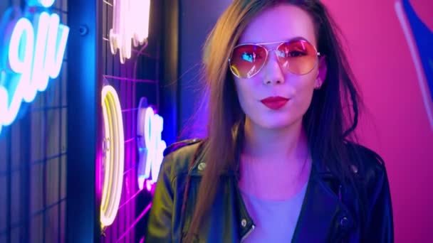 Wanita ras campuran dengan rambut gimbal dan kacamata berpose dekat dinding neon bercahaya dalam gelap — Stok Video