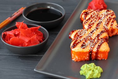 Ahşap masada, siyah tabakta servis edilen fırında suşi rulosu.