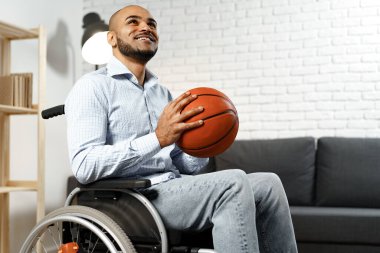 Tekerlekli sandalyede elinde basketbol topu tutan ve gülümseyen mutlu genç adam.