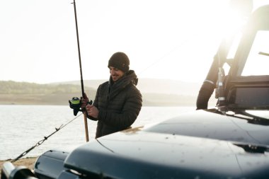 Genç balıkçı arabasının yanında duruyor ve elinde balık ekipmanları var.