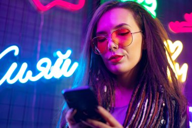Moda kızı gece kulübünde neon tabelaların yanında dururken akıllı telefonunu kullanıyor.