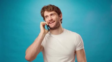 Beyaz gömlekli genç adam telefonda mavi arka planda neşeli bir şekilde konuşuyor.