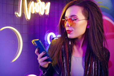 Moda kızı gece kulübünde neon tabelaların yanında dururken akıllı telefonunu kullanıyor.
