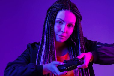 Uzun örgülü genç bir kadın koyu mor arka planda konsol joystick tutuyor.