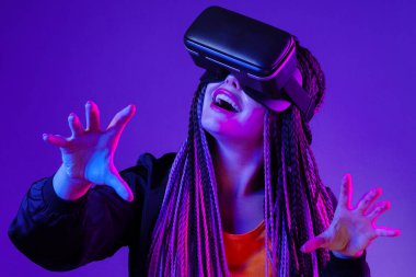 Afro örgülü genç bir kadın koyu mor neon arka planda VR gözlük kullanıyor.