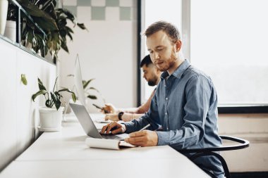 Genç işadamı, modern ofisteki çalışma masasında otururken not defterine notlar alıyor.