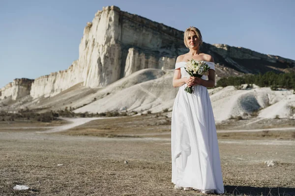 Belle mariée dans une robe de mariée avec un bouquet debout contre le rocher blanc — Photo