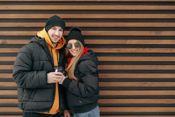 Young couple in winter clothes standing together in a street against wooden wall — Stock Photo, Image