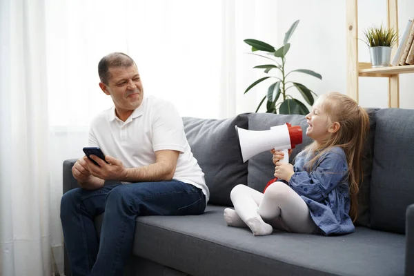 Opa zit op de bank en met behulp van de smartphone, kleine kleindochter schreeuwen door megafoon om hem te stoppen — Stockfoto