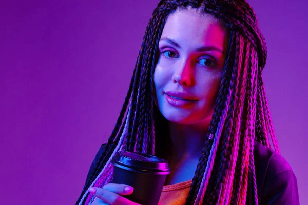 Portrait of a woman with long afro braids holding a cup of coffee against purple neon background — Stock Photo, Image