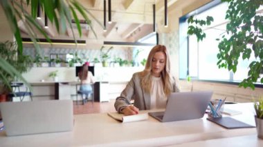 Modern açık uzay ofisindeki masada resmi takım elbiseli genç bir iş kadını çalışıyor.