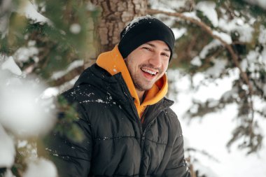 Kapüşonlu ve ceketli genç adam karlı bir parkta duruyor ve gülümsüyor.