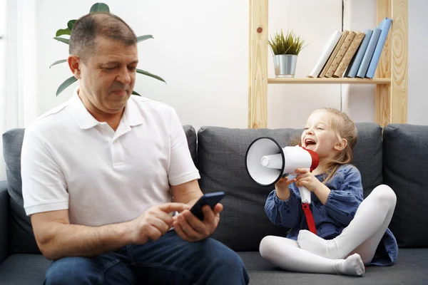 Grandpa sitting on sofa and using smartphone, little granddaughter shouting through megaphone to him to stop — Stock Photo, Image