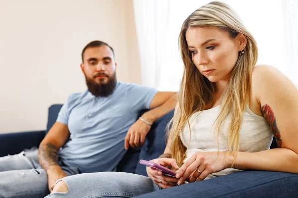 Homem suspeito olhando para a tela do smartphone de suas namoradas enquanto elas se sentam no sofá — Fotografia de Stock