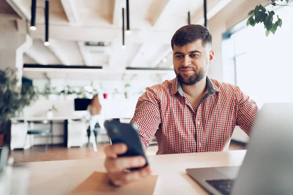 Junger Mann im lässigen Hemd mit Smartphone im Büro, während er am Schreibtisch sitzt — Stockfoto