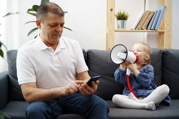 Grandpa sitting on sofa and using smartphone, little granddaughter shouting through megaphone to him to stop — Stock Photo, Image