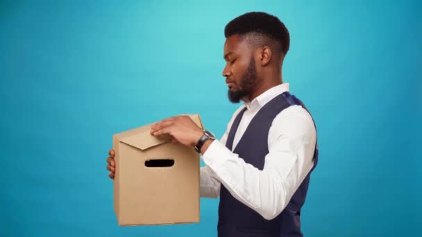 Молодий африканський чоловік відкриває картонну коробку, не любить те, що в ній на синьому фоні — стокове відео