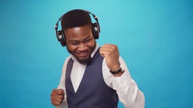 Neşeli Afrikalı adam kulaklıkla müzik dinliyor ve mavi arka planda dans ediyor.