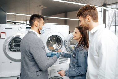 Hipermarkette tıbbi maske takan satıcı müşterilerine yeni bir çamaşır makinesi gösteriyor.