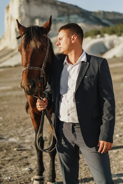 Jovem vestido com um terno preto clássico de pé com um cavalo perto da montanha — Fotografia de Stock