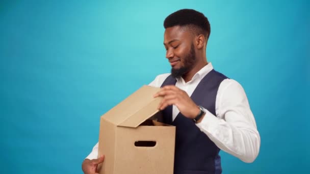 Jonge Afro-Amerikaanse man opent kartonnen doos, houdt van wat erin zit, blauwe achtergrond — Stockvideo