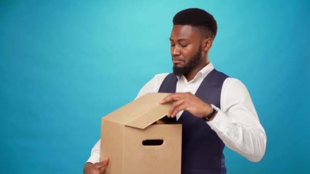 Genç Afrikalı bir adam, mavi arka plandaki kutudan hoşlanmayıp, karton kutuyu açıyor. — Stok video