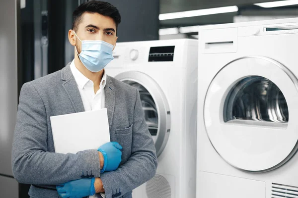 Asistente de tienda masculino en máscara médica demuestra un nuevo modelo de lavadora en el hipermercado — Foto de Stock
