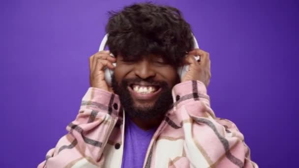 楽しい笑顔アフリカ系アメリカ人の男は、紫の背景にヘッドフォンで音楽を聴く — ストック動画