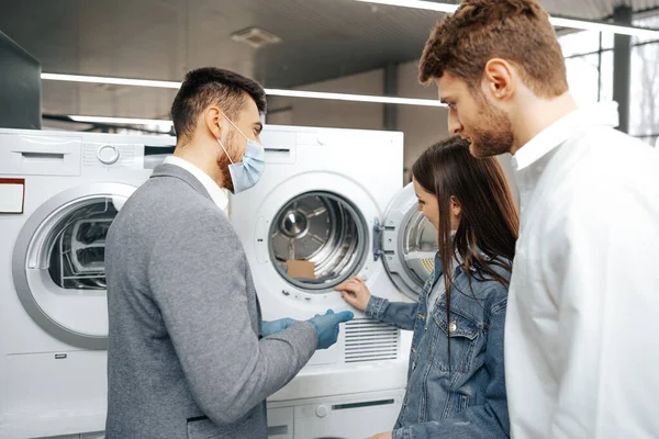 Hipermarkette tıbbi maske takan satıcı müşterilerine yeni bir çamaşır makinesi gösteriyor.