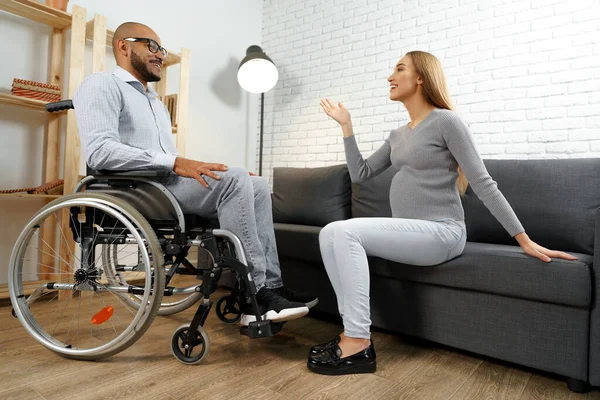 車椅子でアフリカ系アメリカ人の男性が妊娠中の妻とリビングで話しています — ストック写真